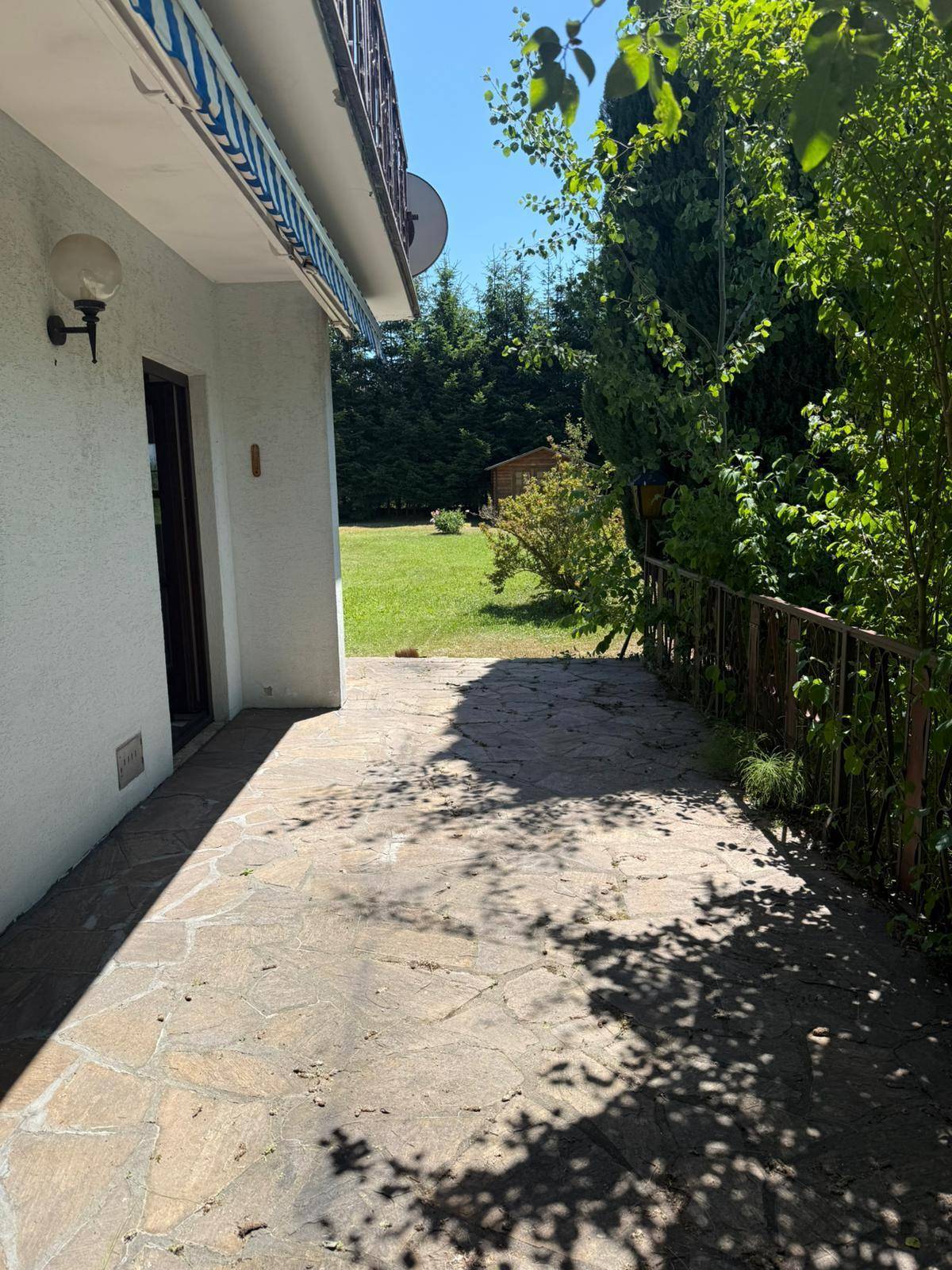 Seitliche Terrasse mit Steinplatten und Metallgeländer, umgeben von viel Grün und Bäumen.