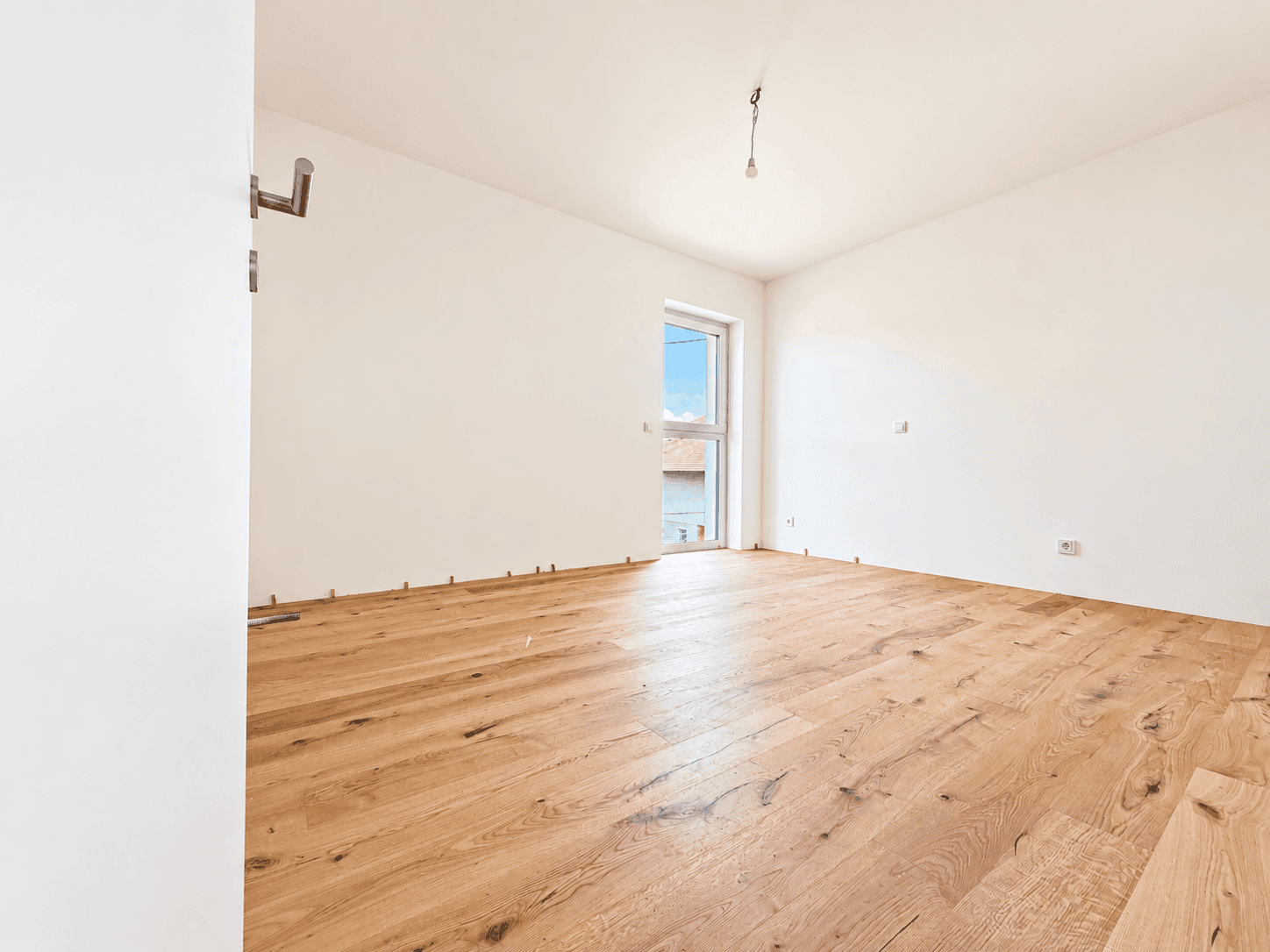 Helles, leeres Zimmer mit Holzboden und einem Fenster, bereit zur individuellen Gestaltung.