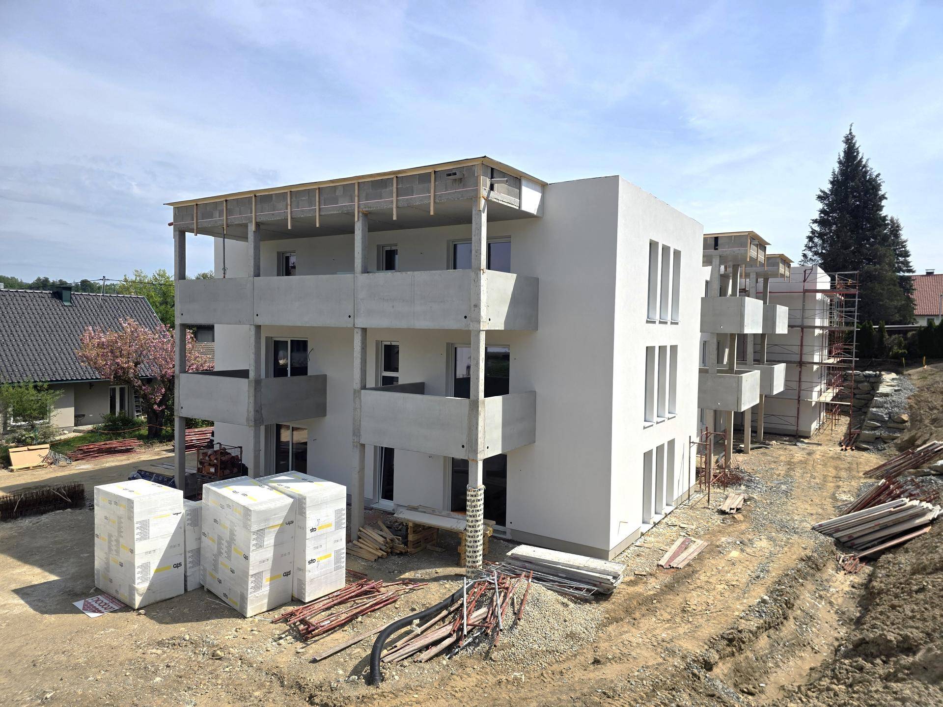Wohngebäude im Bau mit weißer Fassade, großen Fenstern und mehreren Balkonen unter blauem Himmel.