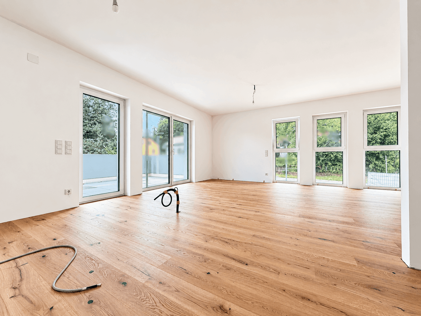Helles, leeres Wohnzimmer mit Holzboden und großen Fenstern, die viel Tageslicht hereinlassen.