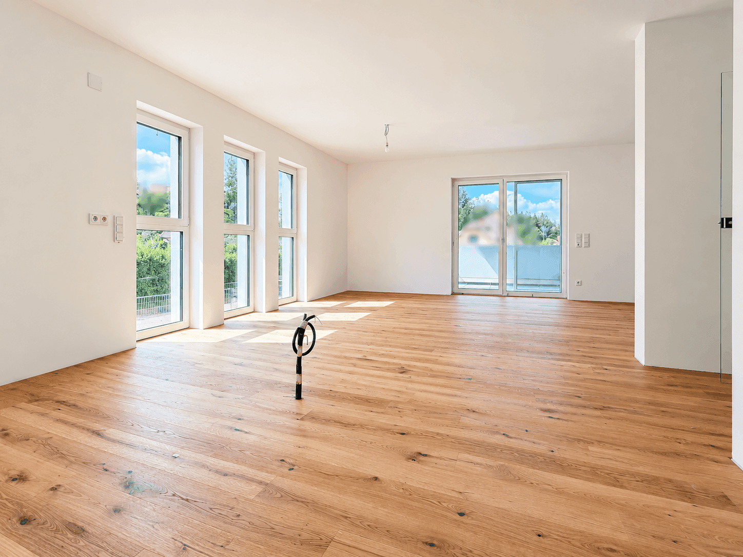 Geräumiger, heller Raum mit Holzboden und großen Fenstern, die Zugang zu einem Außenbereich bieten.