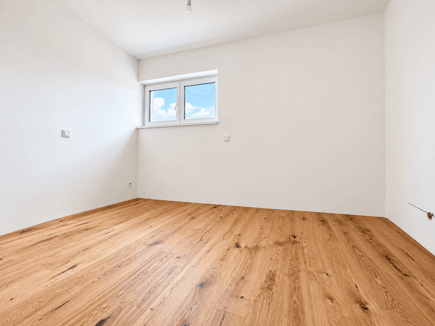 Leeres Zimmer mit Holzboden und einem Fenster, das viel Tageslicht hereinlässt.