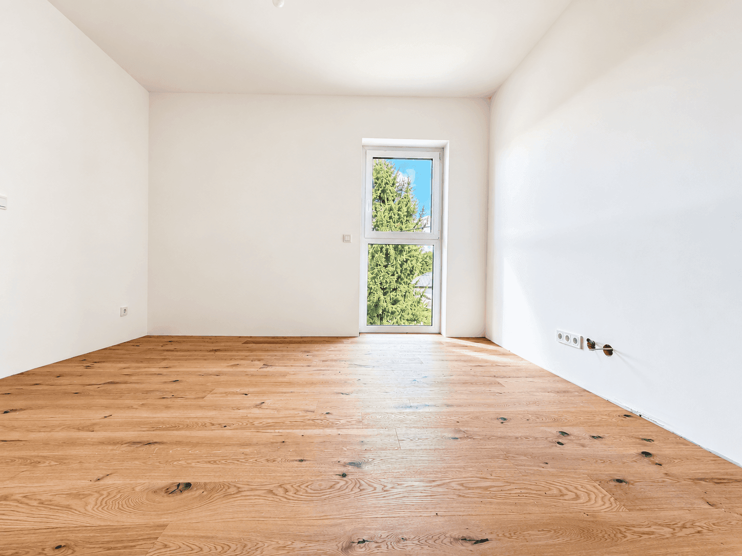 Leeres Zimmer mit Holzboden und einem Fenster, das viel Tageslicht hereinlässt, ideal für ein Kinderzimmer.