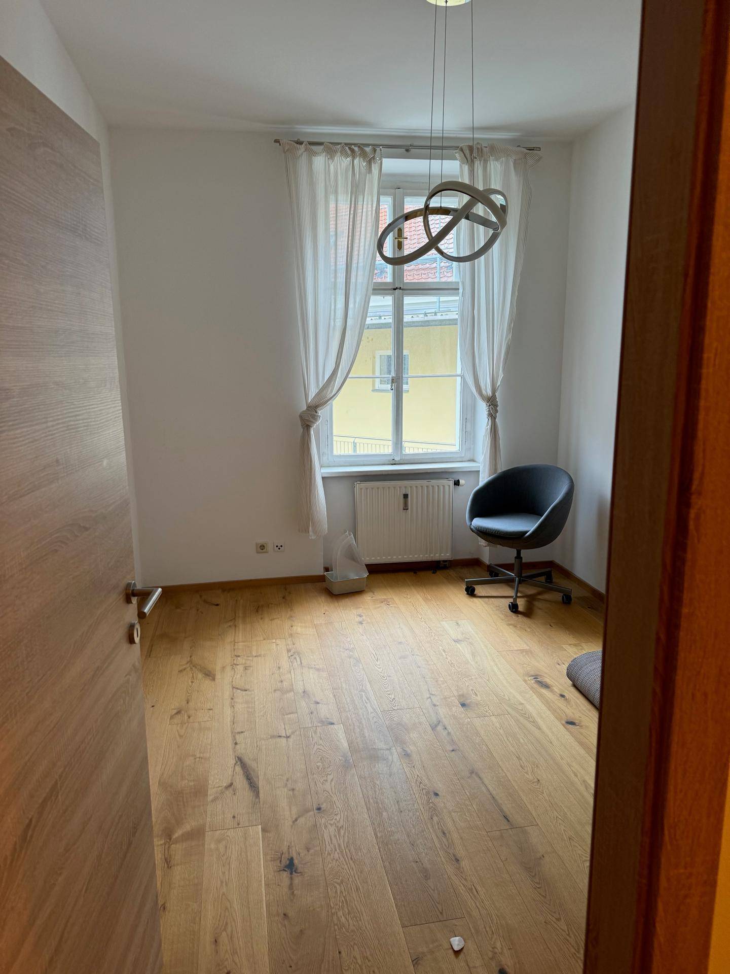 Helles Zimmer mit Holzboden, Fenster und einem Bürostuhl.