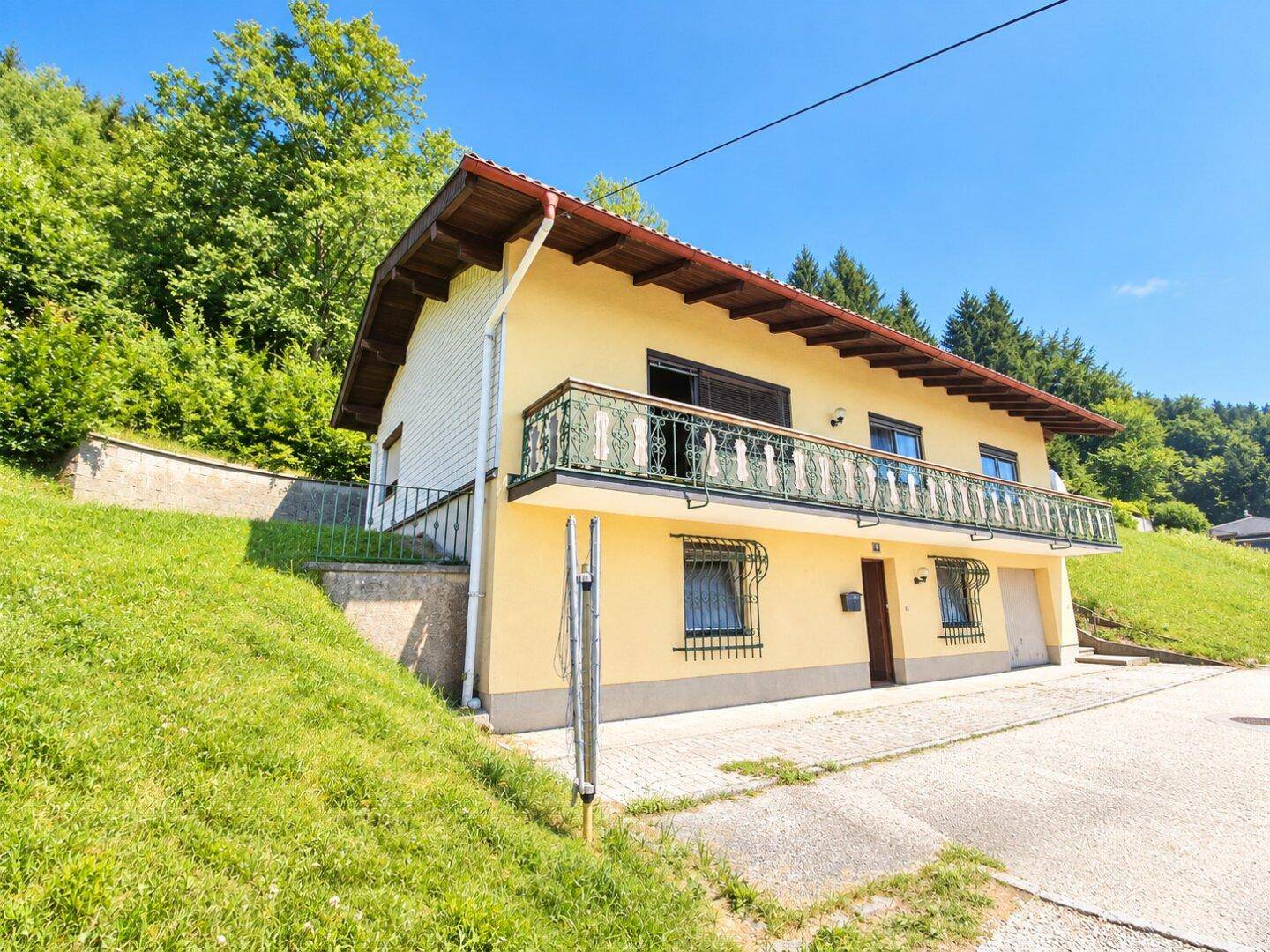 Gelbes Einfamilienhaus mit Balkon und Satteldach, umgeben von grünen Hügeln und Bäumen.
