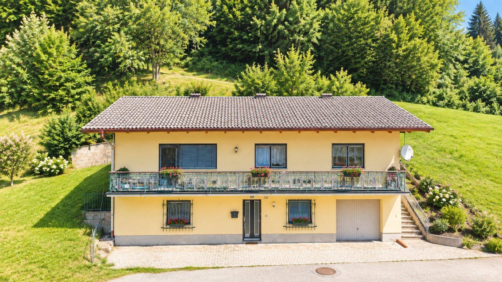 Frontansicht eines gelben Hauses mit Balkon, umgeben von üppiger grüner Landschaft und Bäumen.