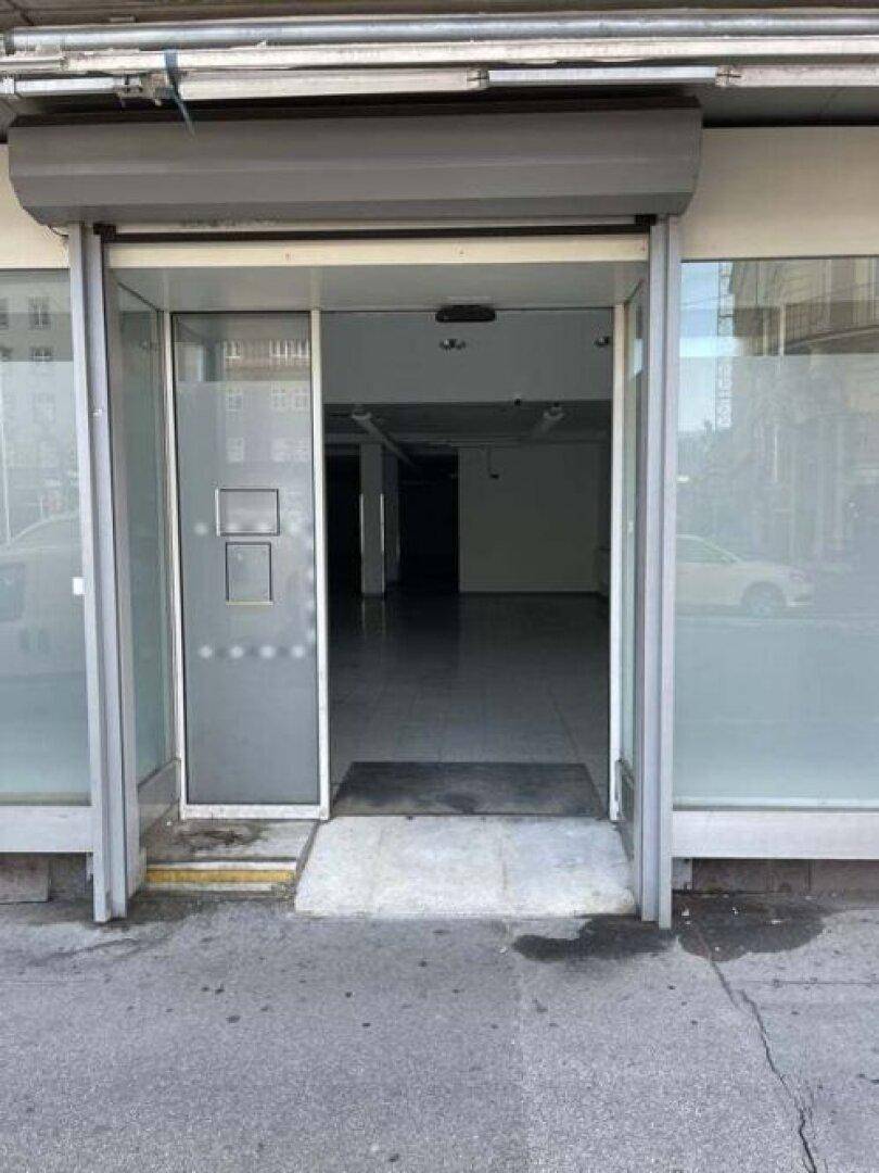 Eingangsbereich des Ladenlokals mit Glastür, Rampe und heruntergelassenem Rollladen.