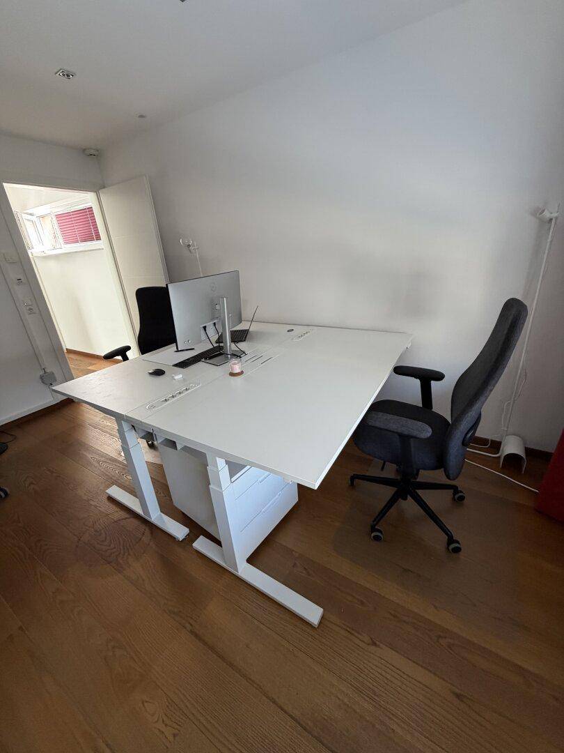 Helles Büro mit einem großen weißen Schreibtisch, ergonomischem Stuhl und Holzboden.
