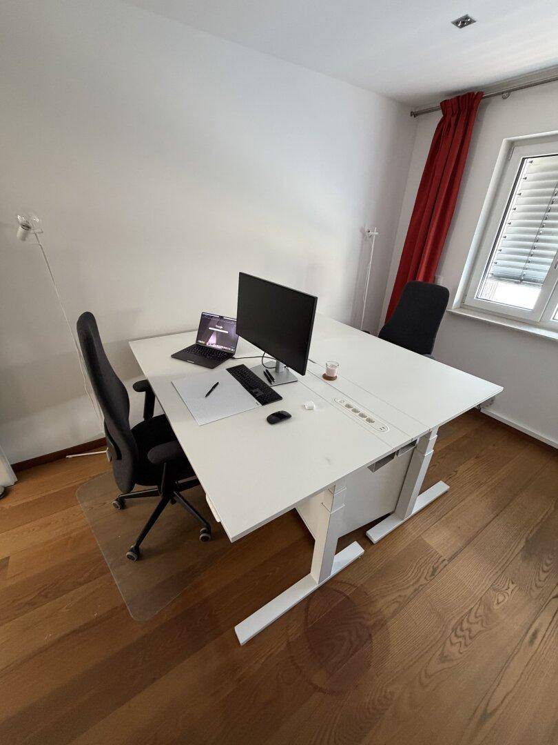 Arbeitszimmer mit einem großen weißen Schreibtisch, Computer und roten Vorhängen an einem Fenster.