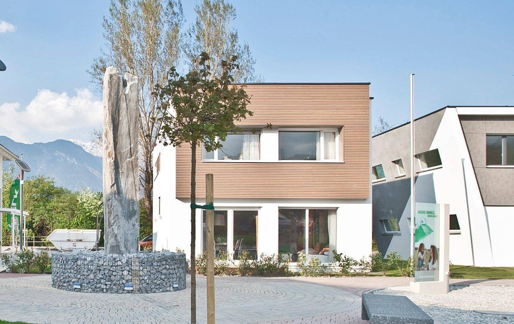 Modernes Haus mit Holzfassade und großen Fenstern, umgeben von einem gepflegten Außenbereich mit Steindenkmal.