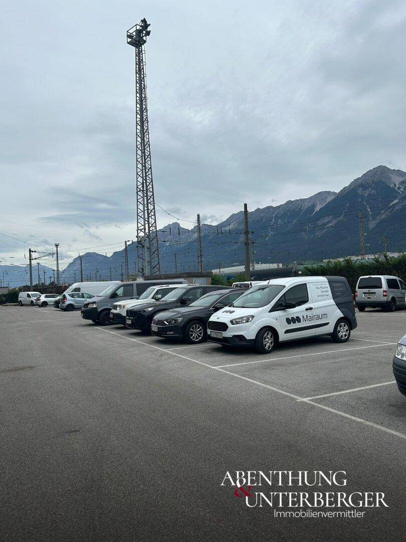 Parkplatz mit mehreren Fahrzeugen, einem hohen Lichtmast und Bergen im Hintergrund.