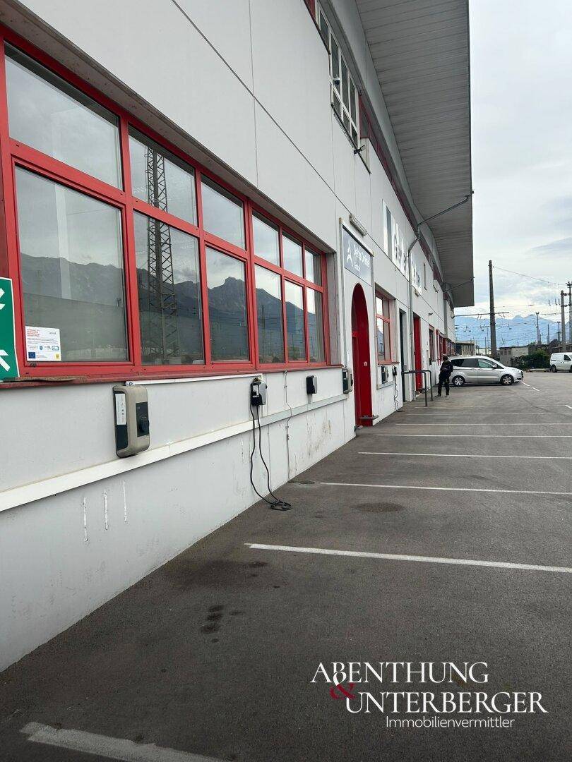 Seitenansicht des Gewerbegebäudes mit Parkplätzen, Bergen im Hintergrund und Bahngleisen.