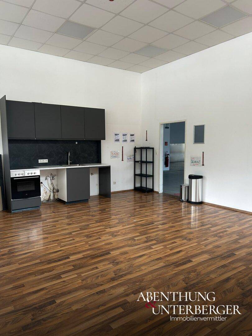 Büro- oder Pausenraum mit Laminatboden und einer kleinen Küchenzeile mit dunklen Schränken und Herd.