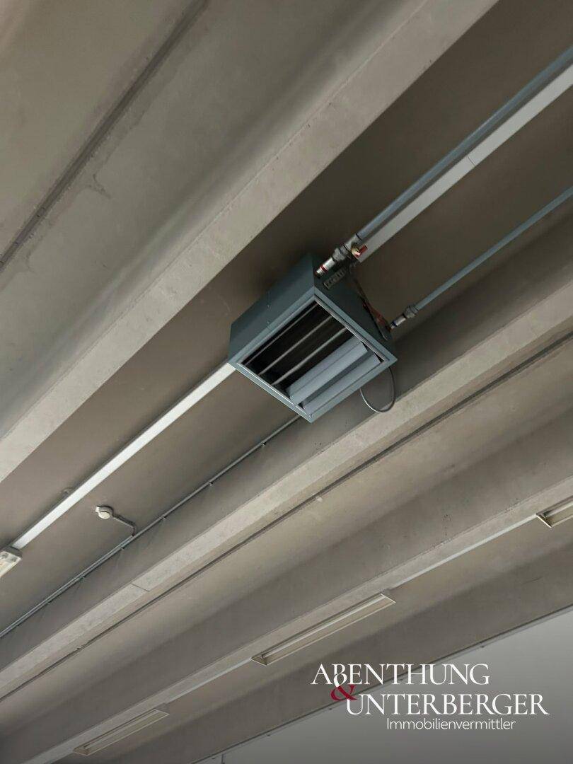 Nahaufnahme der Betondecke mit sichtbaren Rohren und einer Heiz- oder Lüftungseinheit.
