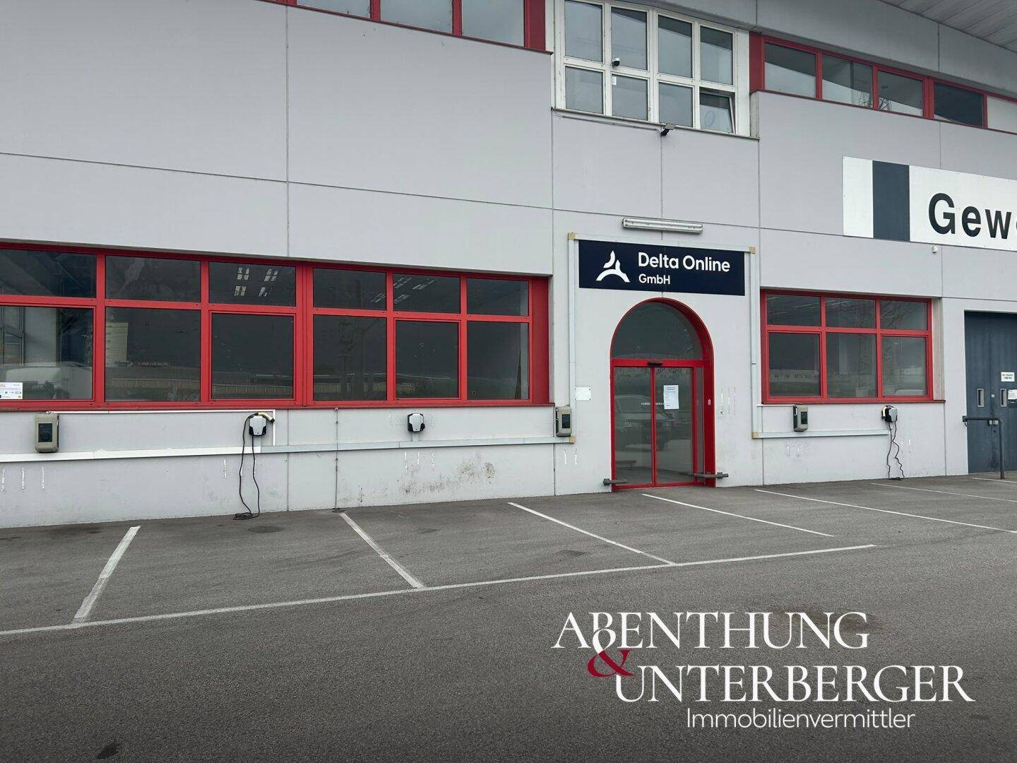 Außenansicht eines Gewerbegebäudes mit roten Fensterrahmen, Parkplätzen und Ladestationen für Elektrofahrzeuge.
