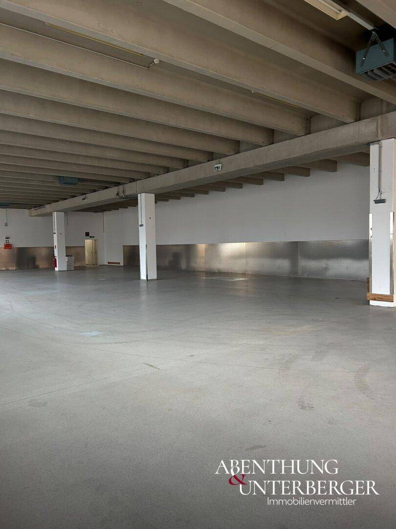 Große, leere Industriehalle mit Betonboden, sichtbaren Deckenbalken und tragenden Säulen.