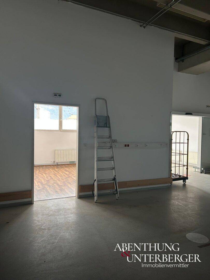 Großer, leerer Raum mit Betonboden und Blick in einen angrenzenden Raum mit Laminatboden.