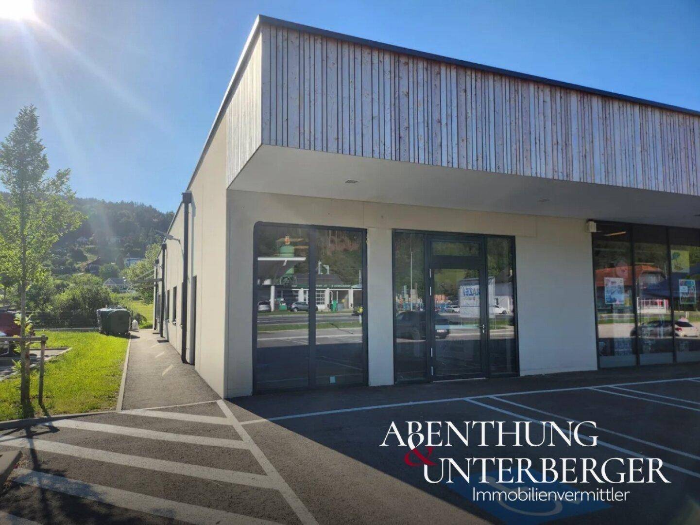Moderne Gewerbefassade mit Holzverkleidung und großen Glasfronten, umgeben von einem Parkplatz und grünen Hügeln.