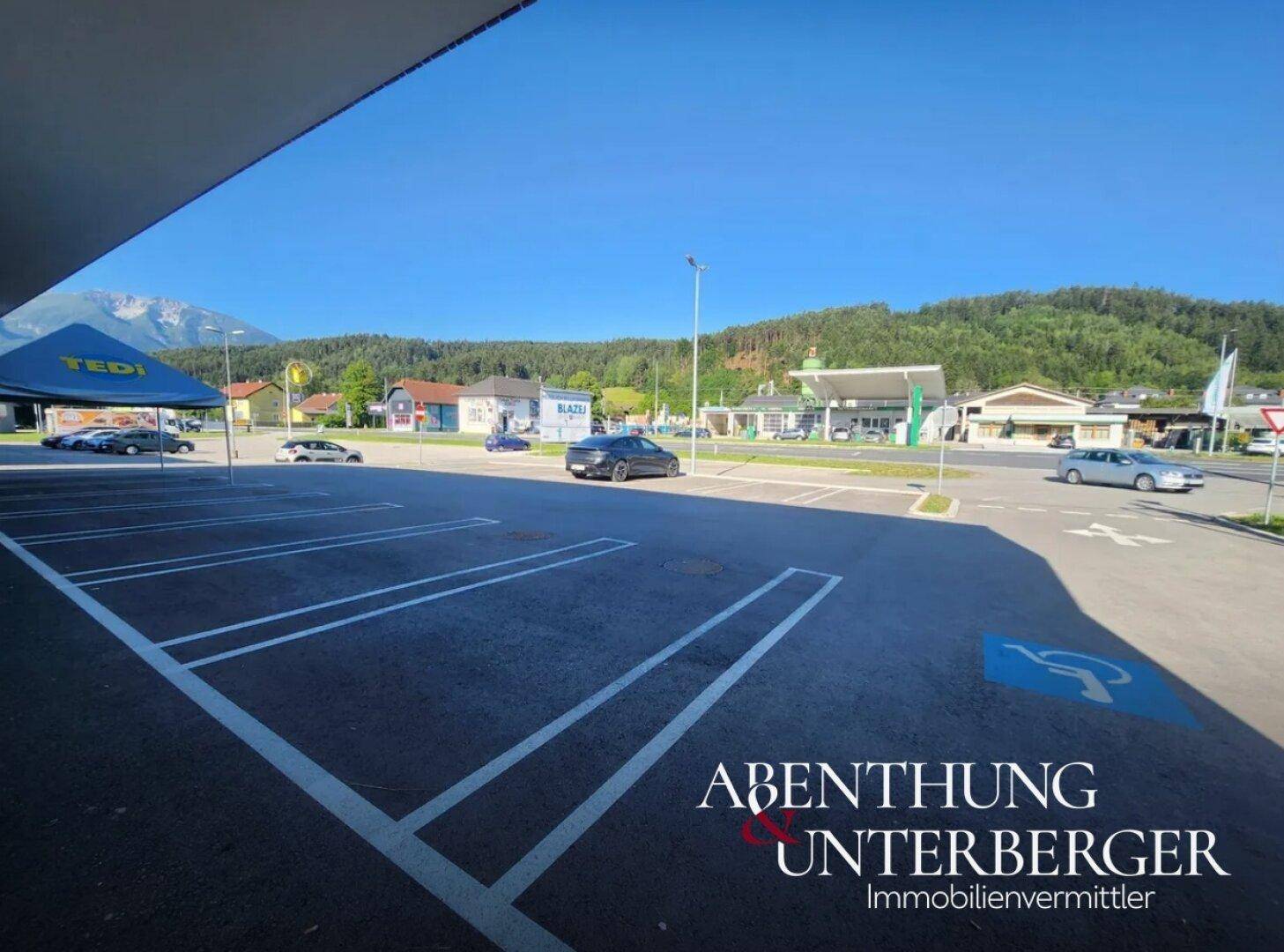 Großer Parkplatz mit markierten Stellplätzen vor einem Gewerbegebiet, umgeben von Bergen und einer Straße.