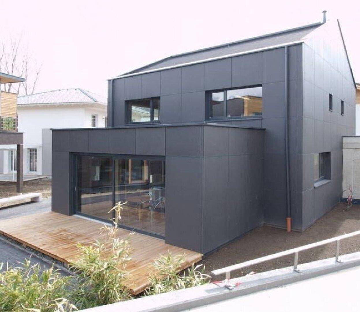 Seitenansicht eines modernen Gebäudes mit Holzterrasse und großen Glasschiebetüren, umgeben von Grünflächen.