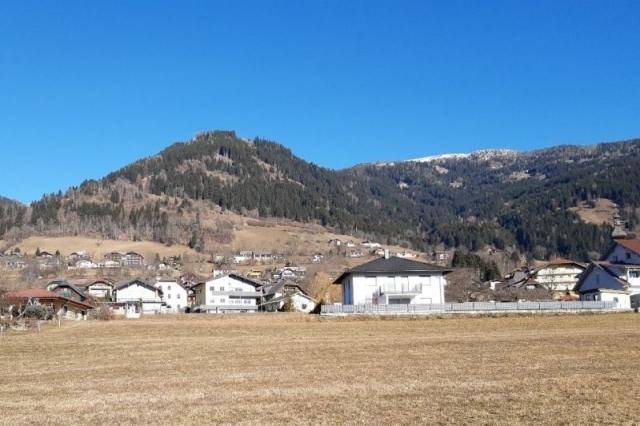 Weitblick über ein trockenes Feld auf eine Ansammlung von Häusern vor einer bewaldeten Bergkette.