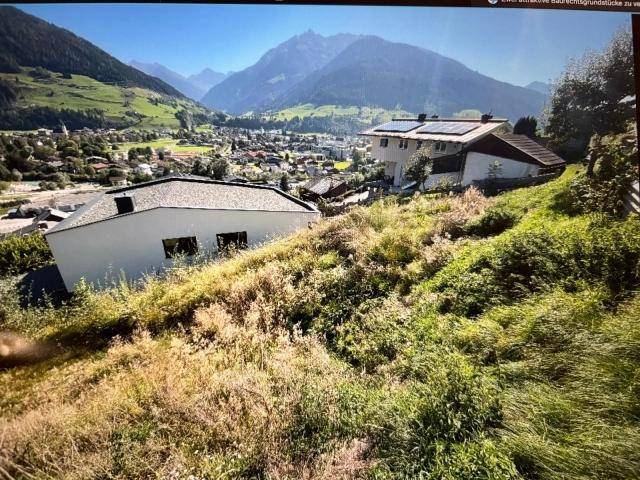 Blick von einem grasbewachsenen Hang auf eine Siedlung im Tal mit umliegenden Bergen.