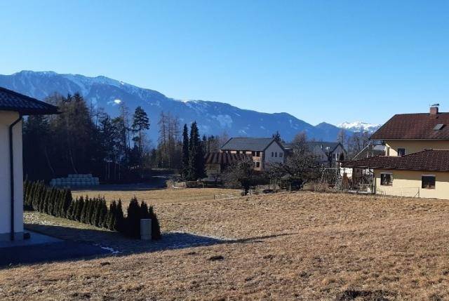 Panoramablick über ein Feld auf eine Wohnsiedlung mit Hecke und schneebedeckten Bergen am Horizont.