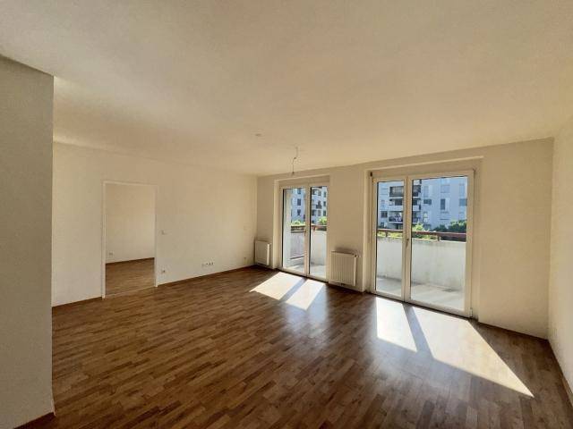 Geräumiges Wohnzimmer mit Holzboden und großen Fenstern, die viel Tageslicht hereinlassen und einen weiteren Raum zeigen.