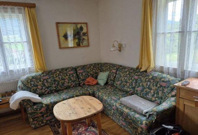 Wohnzimmer mit floral gemustertem Ecksofa, Holztisch und Fenstern mit Blick ins Grüne.