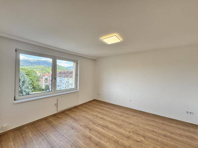 Lichtdurchflutetes Zimmer mit hellem Laminatboden und Ausblick auf die umliegende Berglandschaft und Gebäude.