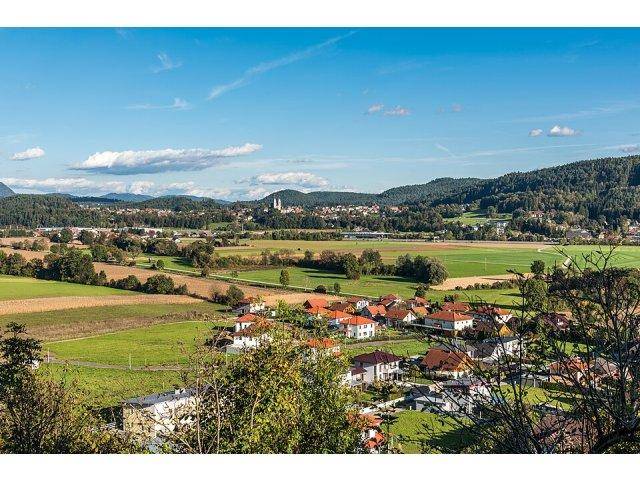 Panoramablick über eine ländliche Landschaft mit einem Dorf, Feldern und Bergen im Hintergrund.