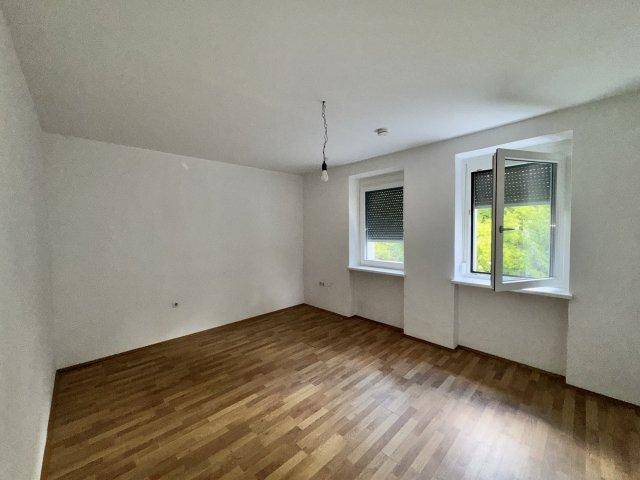Lichtdurchfluteter Raum mit Holzboden und geöffnetem Fenster, das einen Blick ins Grüne bietet.