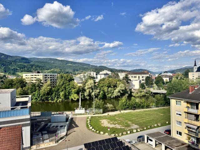 Panoramablick über eine Stadt mit Fluss, grünen Ufern und bewaldeten Bergen im Hintergrund unter blauem Himmel.