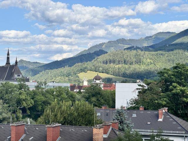 Malerischer Ausblick auf bewaldete Berge, Dächer und eine Kirchturmspitze unter blauem Himmel.