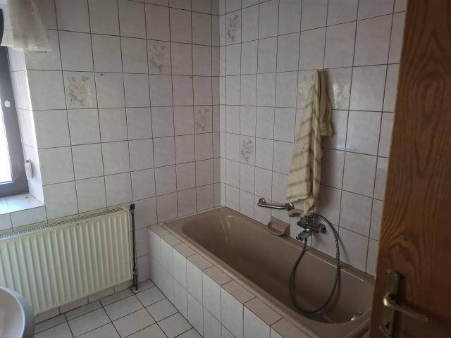 Badezimmer mit Badewanne, hellen Fliesen, Fenster und Haltegriffen im 80er-Jahre-Stil.