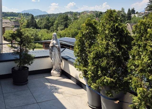 Geräumige Terrasse mit Fliesenboden, Pflanzen und einer Statue, umgeben von grüner Landschaft.