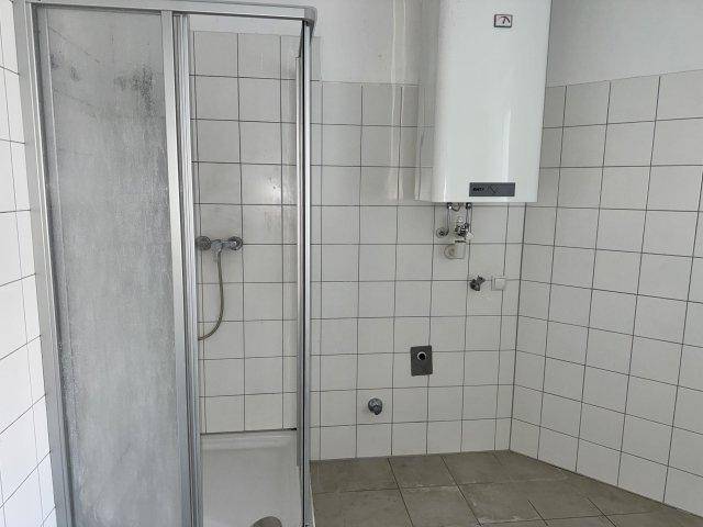 Badezimmer mit Duschkabine und Wandboiler, ausgestattet mit weißen Fliesen und einfacher Einrichtung.