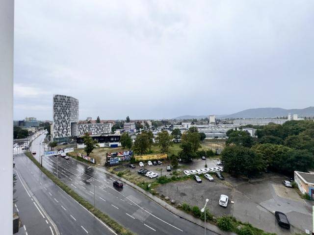 Weitläufiger Blick über die Stadt mit Straßen, Parkplätzen und grünen Hügeln im Hintergrund.