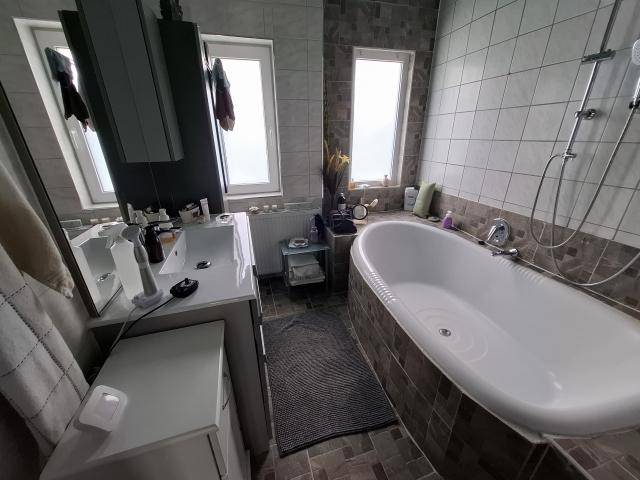 Badezimmer mit Badewanne, Waschtisch, zwei Fenstern und hellen Fliesen an Wänden und Boden.