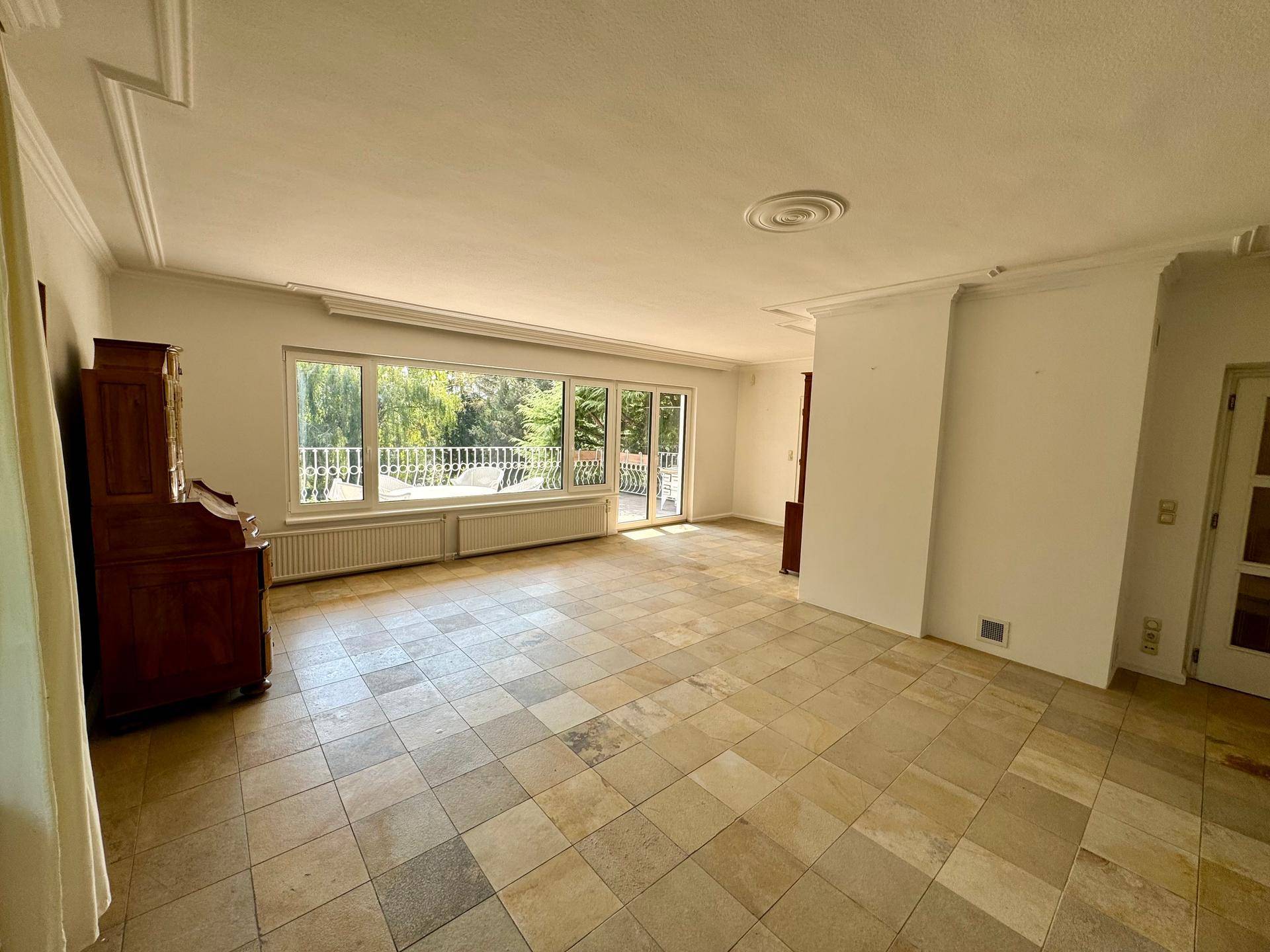 Großzügiges Wohnzimmer mit Natursteinfliesen, Stuckdecke und Zugang zur Terrasse.