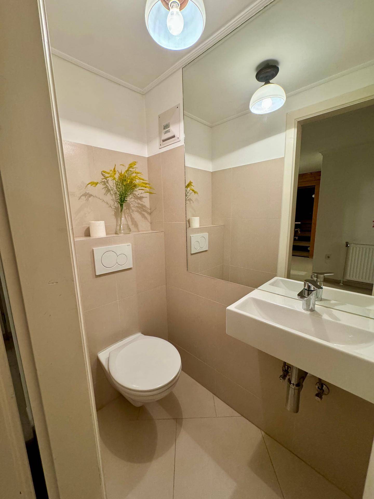 Modernes Gäste-WC mit Wand-WC, kleinem Waschbecken und beigen Fliesen an Wänden und Boden.