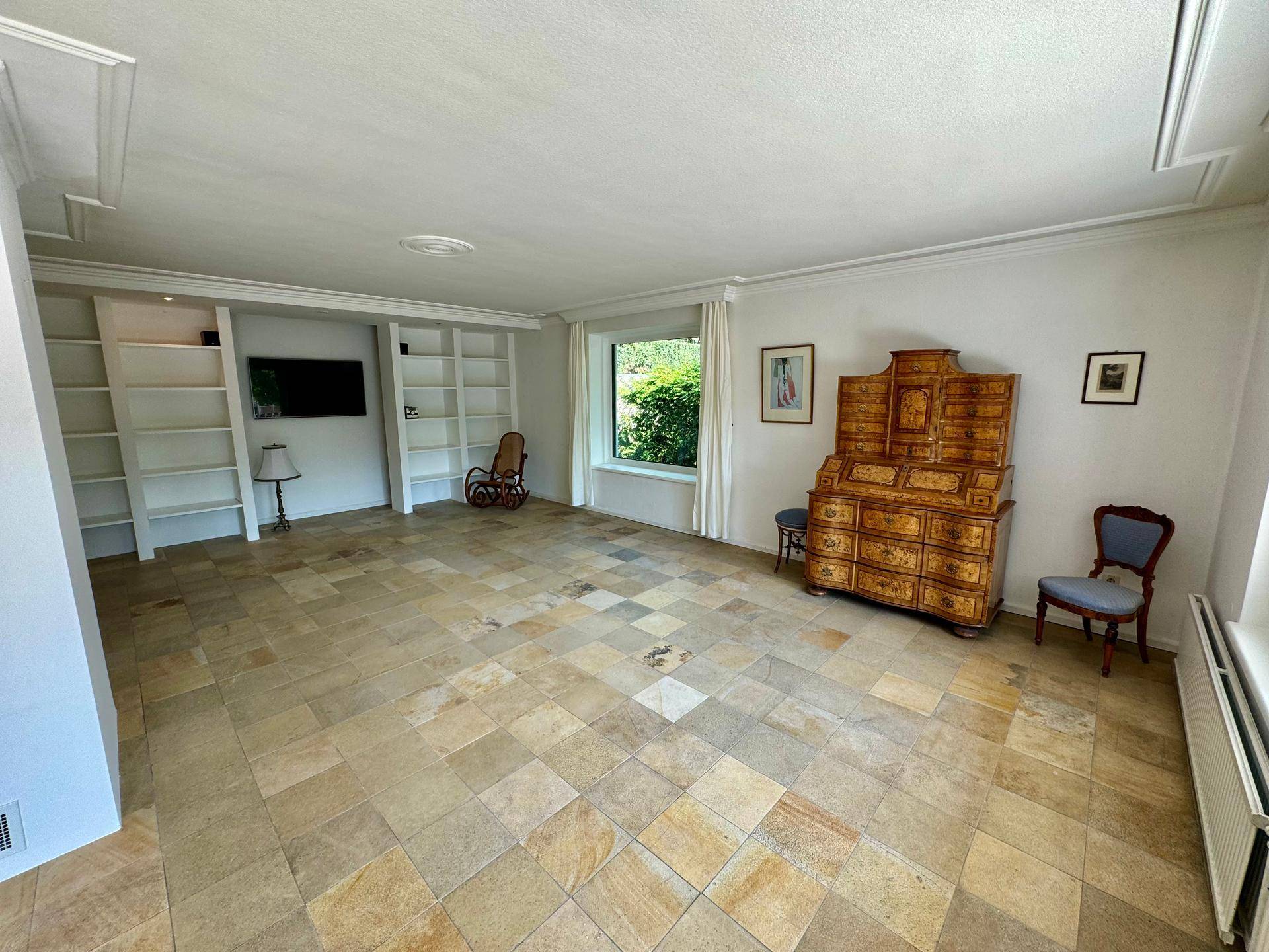 Helles Wohnzimmer mit Natursteinfliesen, Einbauregalen und Blick ins Grüne.