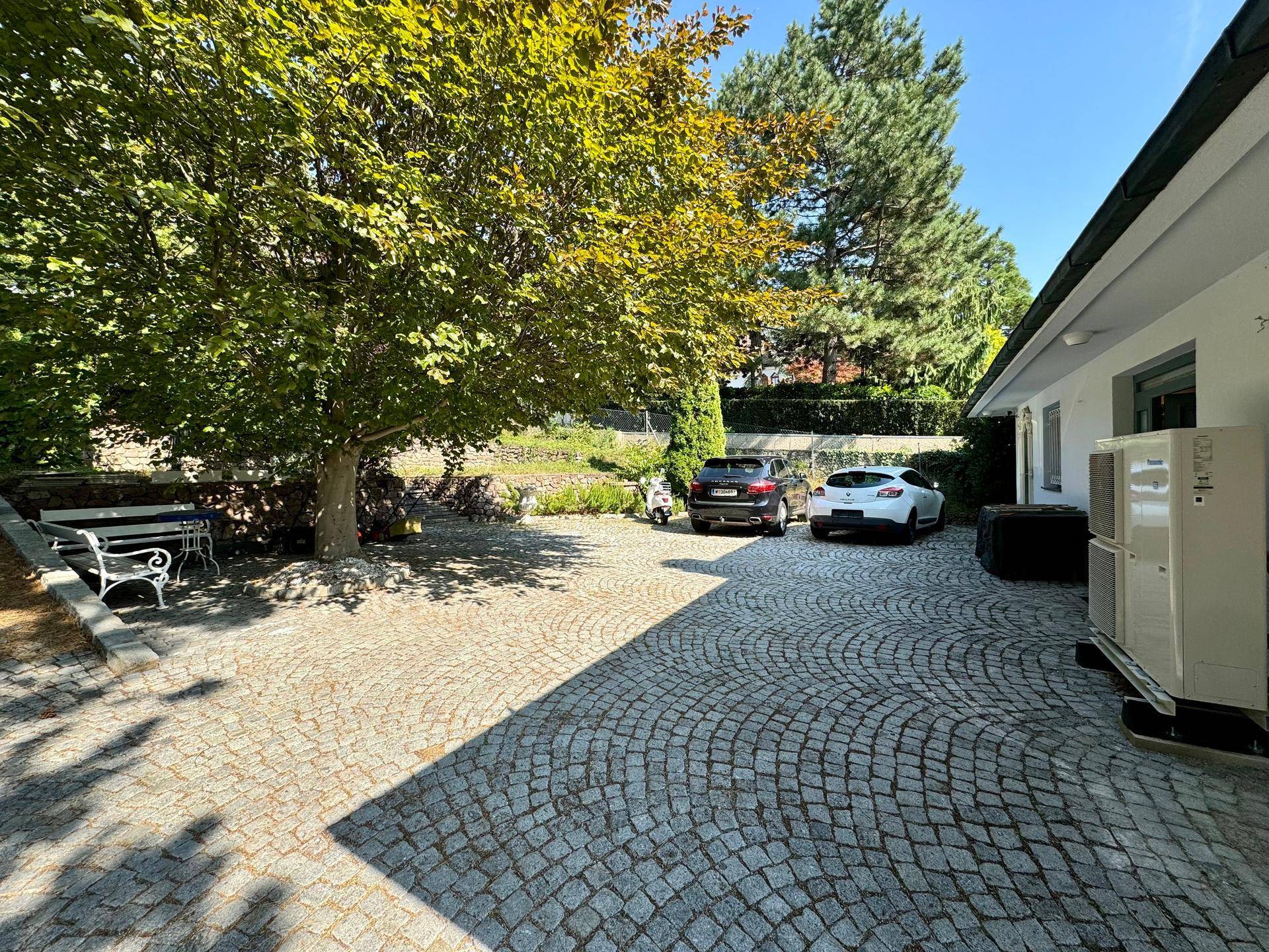 Geräumiger gepflasterter Parkplatz mit Baumbestand, viel Sonne und einer Wärmepumpe.