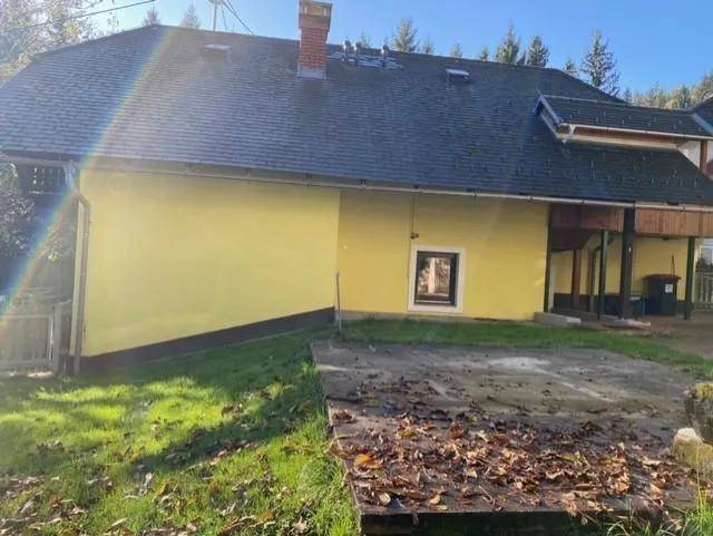 Rückansicht eines Hauses mit gelber Fassade und dunklem Dach, umgeben von Rasen und Bäumen.