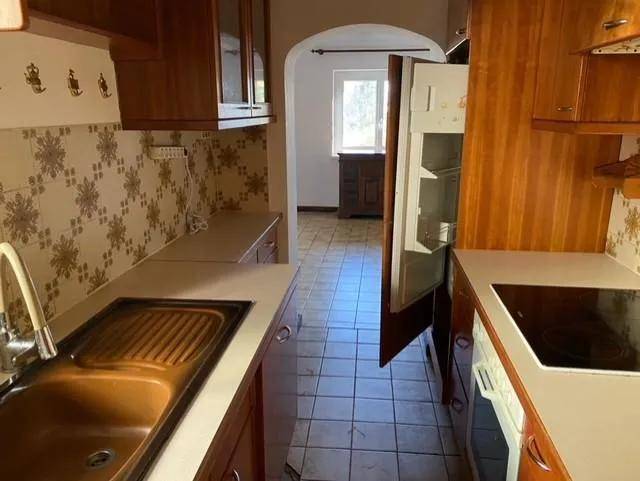Längsansicht der Küche mit Holzmöbeln, Kühlschrank und einem Durchgang zu einem weiteren Raum.