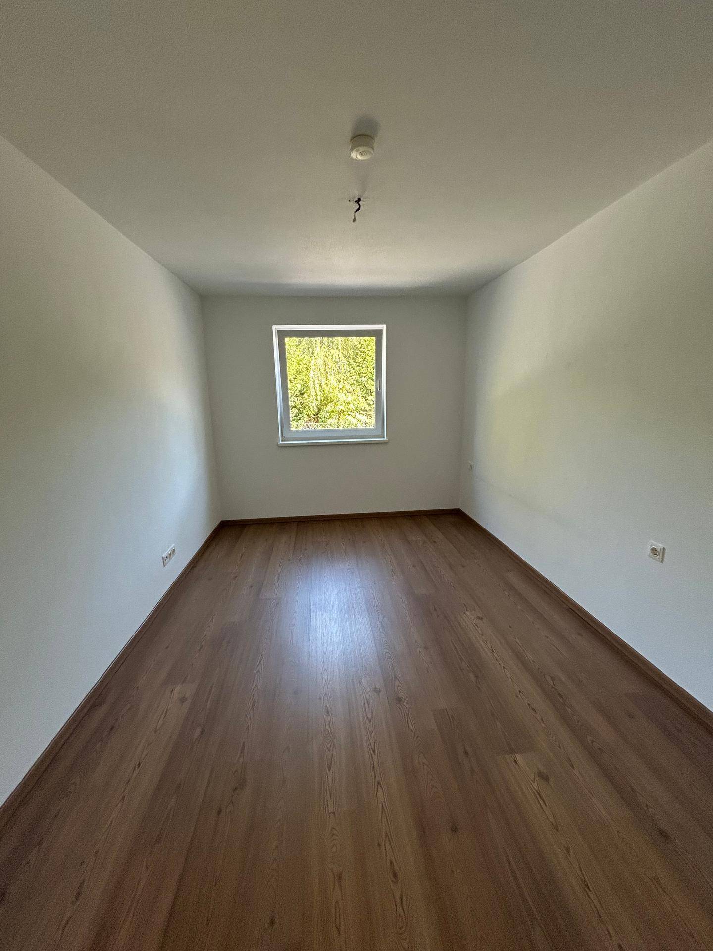 Leeres Zimmer mit Holzboden und einem Fenster, das viel Tageslicht hereinlässt.