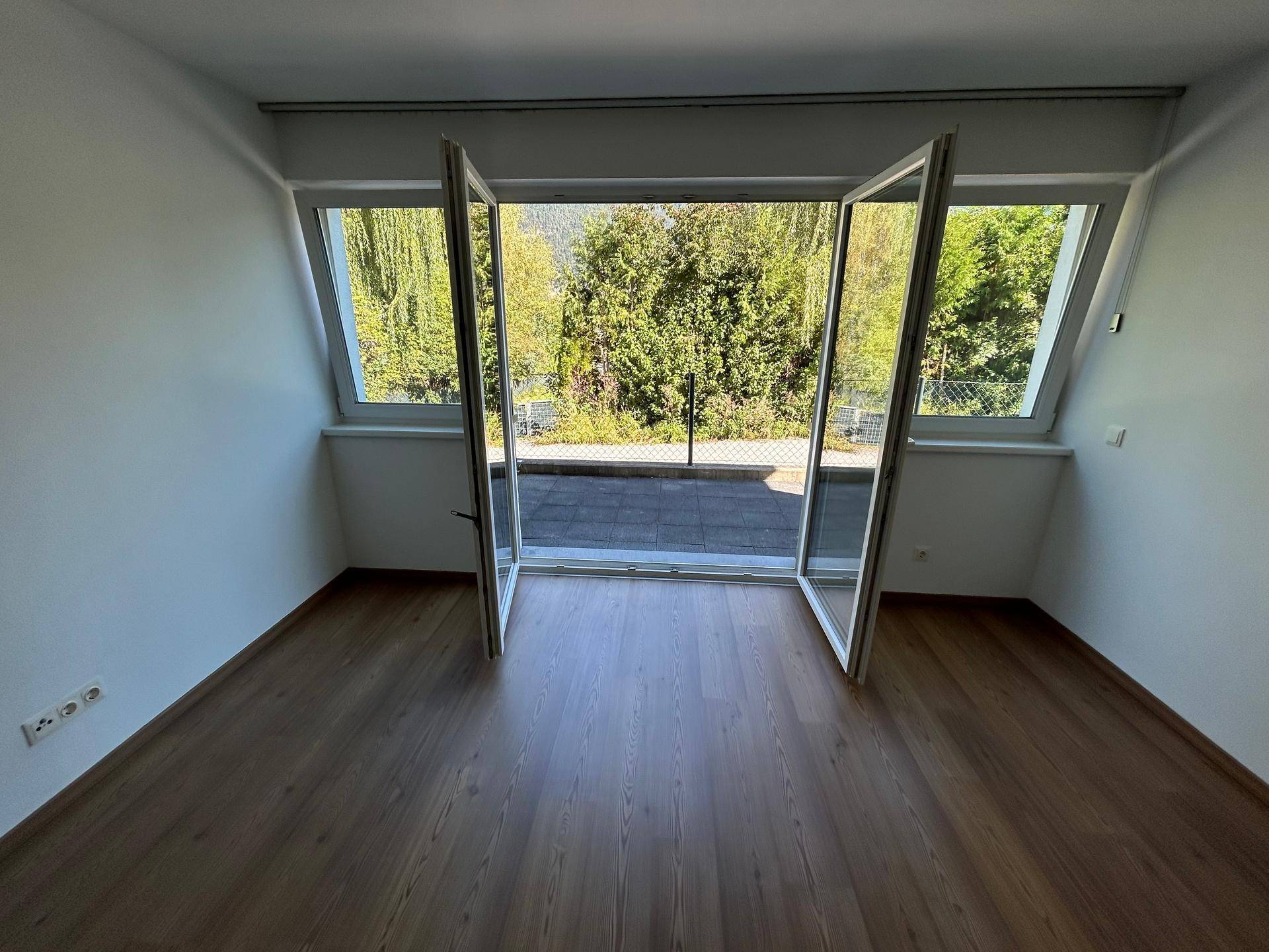 Helles Zimmer mit Holzboden und großen Fenstertüren, die auf einen Balkon führen.