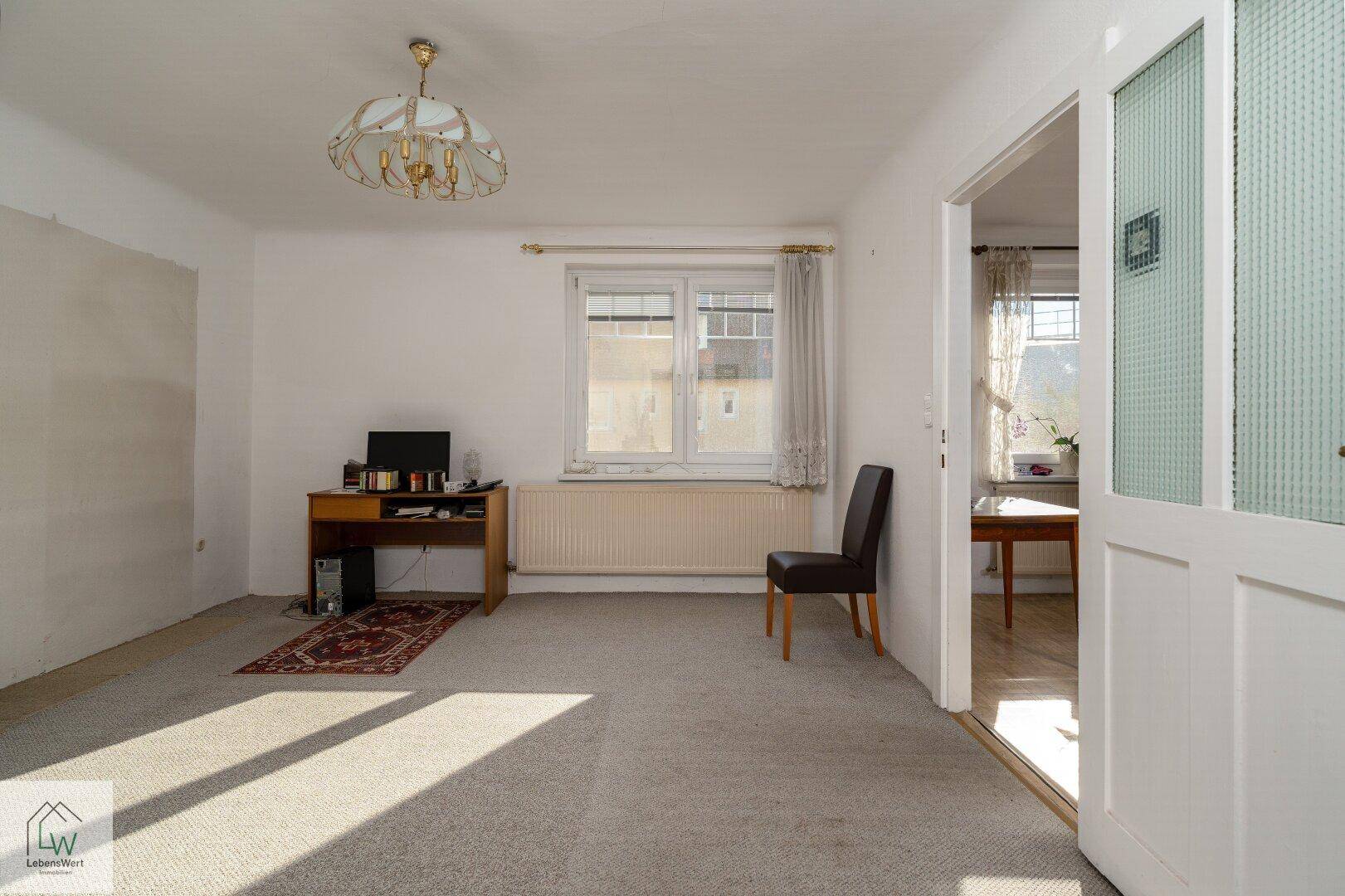 Helles Wohnzimmer mit Teppichboden, Schreibtisch und Blick auf die Umgebung.