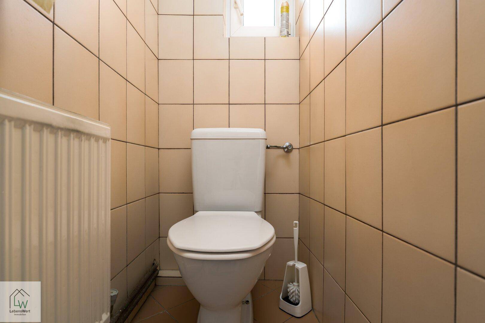 Kleines WC mit beigefarbenen Fliesen, Toilette und Fenster für natürliche Belüftung.