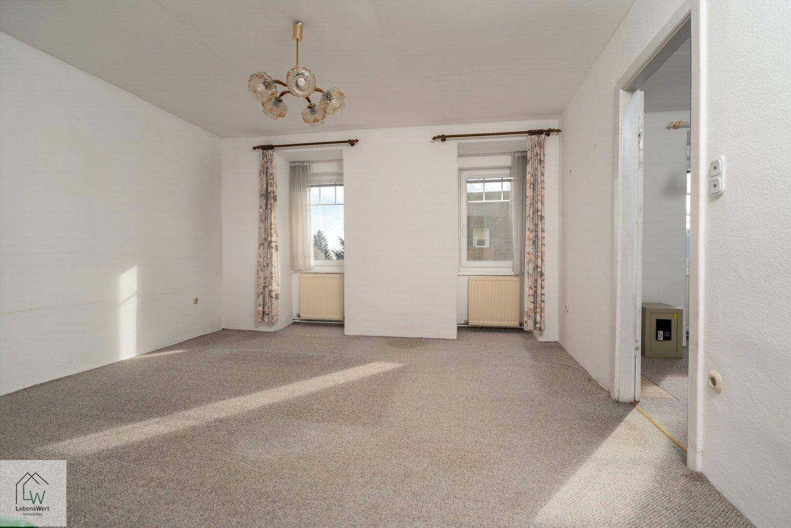 Helles Wohnzimmer mit Teppichboden und zwei Fenstern, die viel Tageslicht hereinlassen.